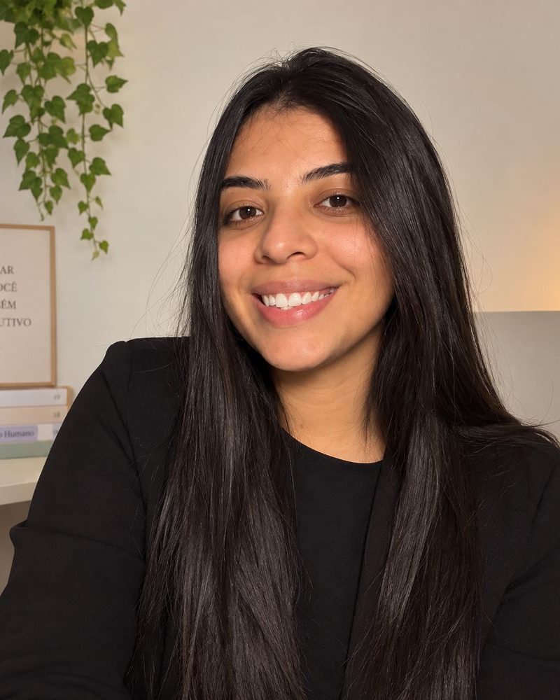 Larissa Lima, psicóloga, sorrindo, de cabelos longos e escuros, em uma sala clara com uma planta e livros ao fundo.