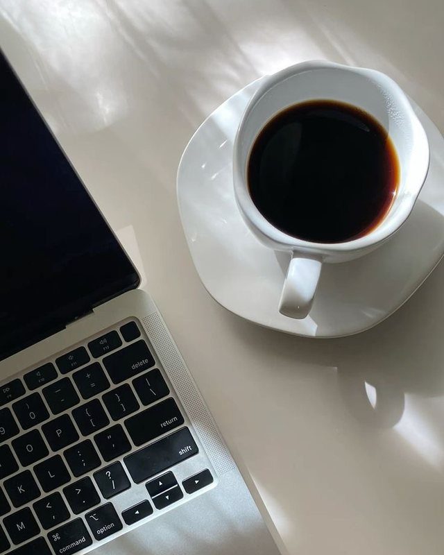 Uma xícara de café ao lado de um notebook aberto, sobre uma mesa clara iluminada pelo sol.